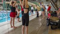 Een groep zwemmers doet onder begeleiding een stretchingsoefening van de triceps naast het zwembad