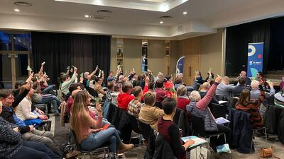 Foto van de algemene vergadering van G-sport Vlaanderen. De aanwezige leden stemmen en steken groene of rode kaartjes in de lucht.